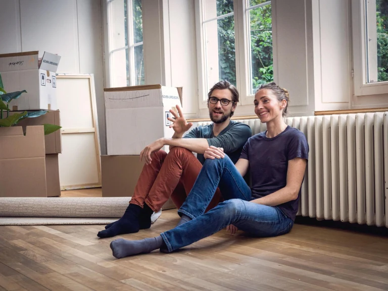 Junges Paar sitzt umgeben von Umzugskarton in einer nicht möblierten Wohnung